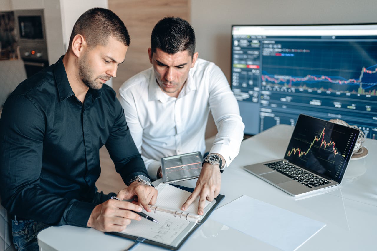 about-01 Two businessmen analyzing financial charts on digital devices in a modern office setting.